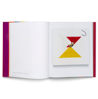 A photo of an open art book on a white surface. The right page displays an abstract geometric painting featuring a red triangle over a yellow triangle, creating an hourglass shape with smaller black and white elements at the center. The left page is mostly blank with a purple inner margin and a caption at the bottom.