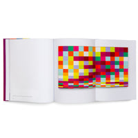 An open art book with a magenta spine, set against a white background. The right-hand pages feature a vibrant, fold-out image of an abstract grid pattern made of multicolored squares and rectangles. The left page is mostly blank with a small caption.