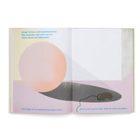 An open children's book with German text, showing a two-page illustration. A large, pale orange sphere casts a dark grey shadow across the bottom of the pages. On the right, inside the shadow, a small, brown, mouse-like creature with a long green tail is sleeping. The background consists of pastel yellow and pink geometric shapes.