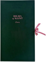 A dark green, cloth-bound book or portfolio cover with the title HILMA af KLINT Flora printed in a light pink font. A matching pink ribbon is tied in a bow on the right side, serving as a closure. The book is set against a plain white background.