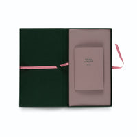 A top-down view of an open, dark green presentation box with a dusty pink interior. Nestled inside on the right is a smaller pink booklet with the text HILMA af KLINT Flora. A pink ribbon closure is untied and visible on both sides of the box.