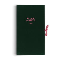A dark green, cloth-bound book titled HILMA af KLINT Flora in bright pink text. The book is shown vertically against a white background, and a pink ribbon tab sticks out from the right side.