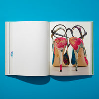 A flat lay of an open book on a vibrant blue background. The left page is blank, while the right page displays a photograph of the back of a pair of colorful, floral-patterned high-heeled slingback shoes. A pair of round, black-framed eyeglasses is resting on the shoes.
