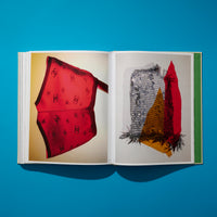 A flat lay, overhead photograph of an open book on a bright blue surface, casting a shadow. The left page shows a sheer red scarf with a black Chanel logo pattern against a yellow background. The right page features a photo of a mixed-media collage with silver chainmail, gold sequins, and red fabric.