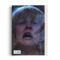 The back cover of a book with an off-white canvas spine. The cover features a grainy, blue-tinted, and blurry close-up of a person's face in distress, with their mouth open. A faint, translucent hand is pressed against the right side of their face. A barcode is in the lower-left corner.