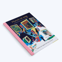 A book with a pink spine, titled Munch ist weg, lies at an angle on a white surface. The dark blue cover features a colorful, busy illustration of a multi-level art gallery where stylized people view famous Edvard Munch paintings, including The Scream.