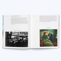 An open book with German text laid flat on a white surface. The left page features a black-and-white photograph of men at an art exhibition. The right page displays a color reproduction of a painting showing a sick person in bed and another figure grieving at their side.