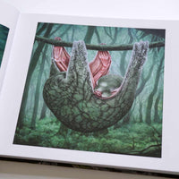 A close-up of an open art book displaying a surreal illustration of a creature resembling a sloth. The animal has a green, textured body and pink, muscular underarms, and it hangs sleeping from a branch in a misty, dark green forest.