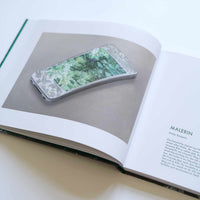 An open art book on a white surface. The left page displays a realistic illustration of a white smartphone with a vibrant green, leafy image on its screen, set against a grey background. The right page contains German text under the heading MALERIN by Walter Benjamin.