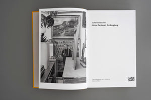 An open art book with a yellow spine lies on a grey surface. The left page features a black-and-white photograph of a person in a room filled with art and objects. The right page is a title page with text, including the names Julia Gaisbacher and Hanne Darboven, and the publisher logo for Hatje Cantz.