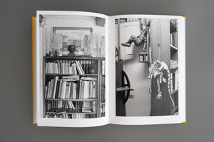 An open book with a yellow cover lies on a grey background, displaying a two-page black and white photograph. The image shows a cluttered room with a bookshelf full of books, art, and sculptures, including a figure hanging upside down and a small dinosaur skeleton dangling from a cabinet.