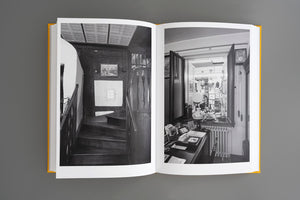 A yellow-spined book is open to a two-page spread of black and white photographs. The left page shows a dark, narrow wooden staircase in a rustic interior. The right page shows a cluttered desk with a lamp and framed photos next to an open window that looks into another room filled with objects.