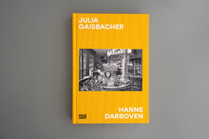 A book with a bright yellow textured cover, titled Julia Gaisbacher, Hanne Darboven, rests on a gray surface. The cover features a black and white photograph of a cluttered room filled with various objects, including display cases, shelves, and a model of a skyscraper. The publisher's logo, Hatje Cantz, is printed at the bottom.